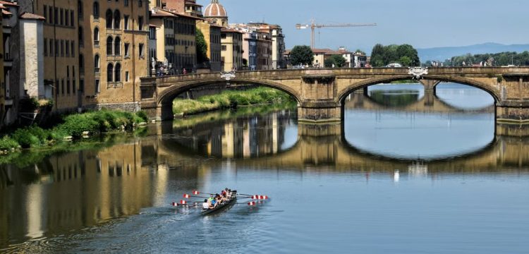 Rowers glide under a historical bridge in Florence, reflected in the river.