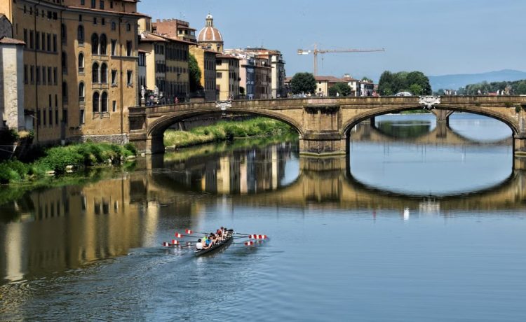 Rowers glide under a historical bridge in Florence, reflected in the river.